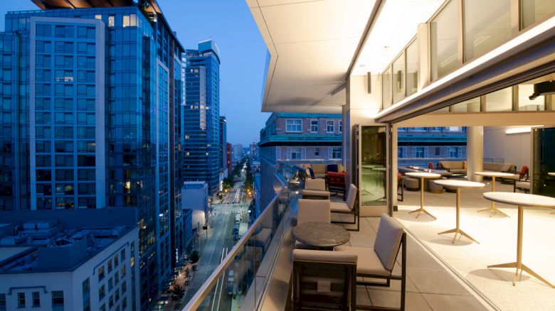 A modern balcony view overlooking a cityscape at dusk, with sleek furnishings and high-rise buildings lining the street below.