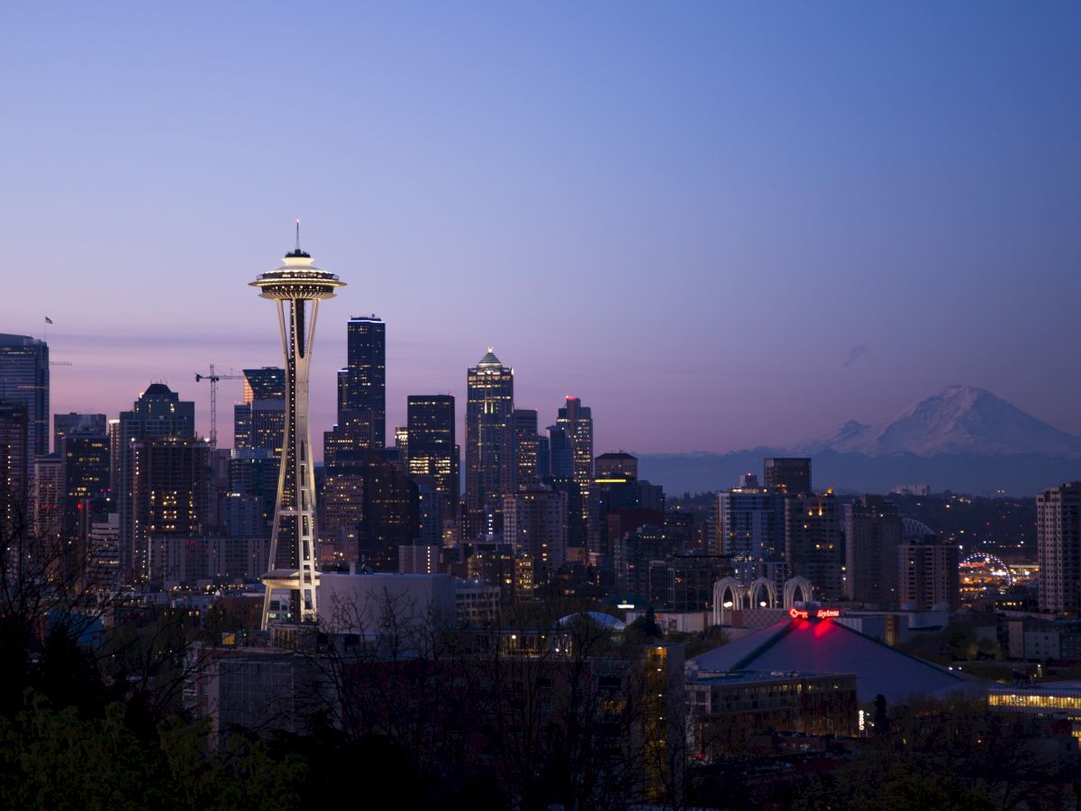 Seattle skyline at dusk with the Space Needle illuminated against a purple sky, and Mount Rainier visible in the distance.