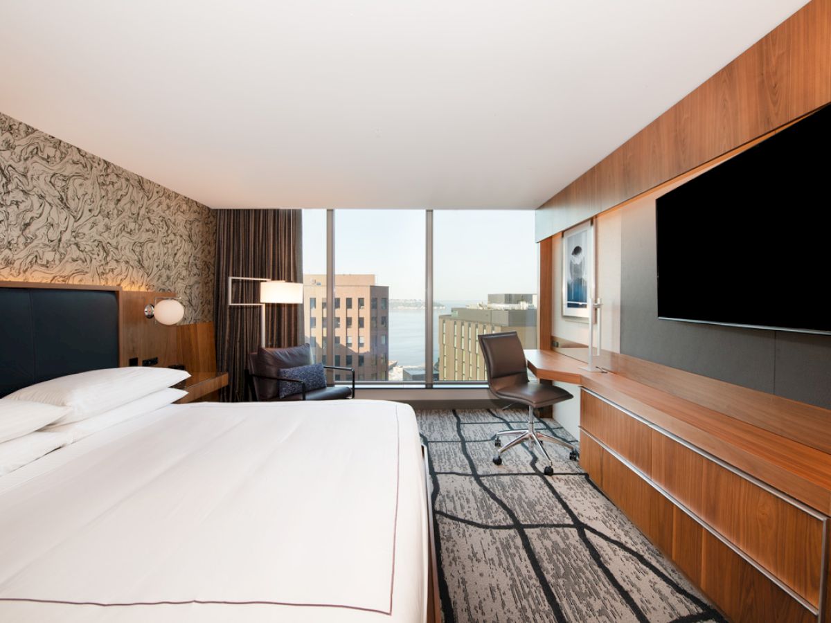 A modern hotel room with a large bed, desk, chair, wall-mounted TV, and window with a cityscape view.