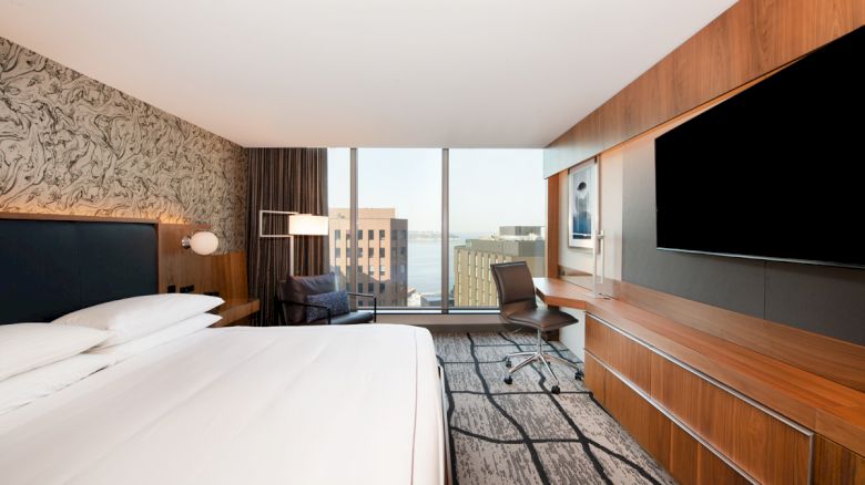 A modern hotel room with a large bed, desk, chair, wall-mounted TV, and window with a cityscape view.