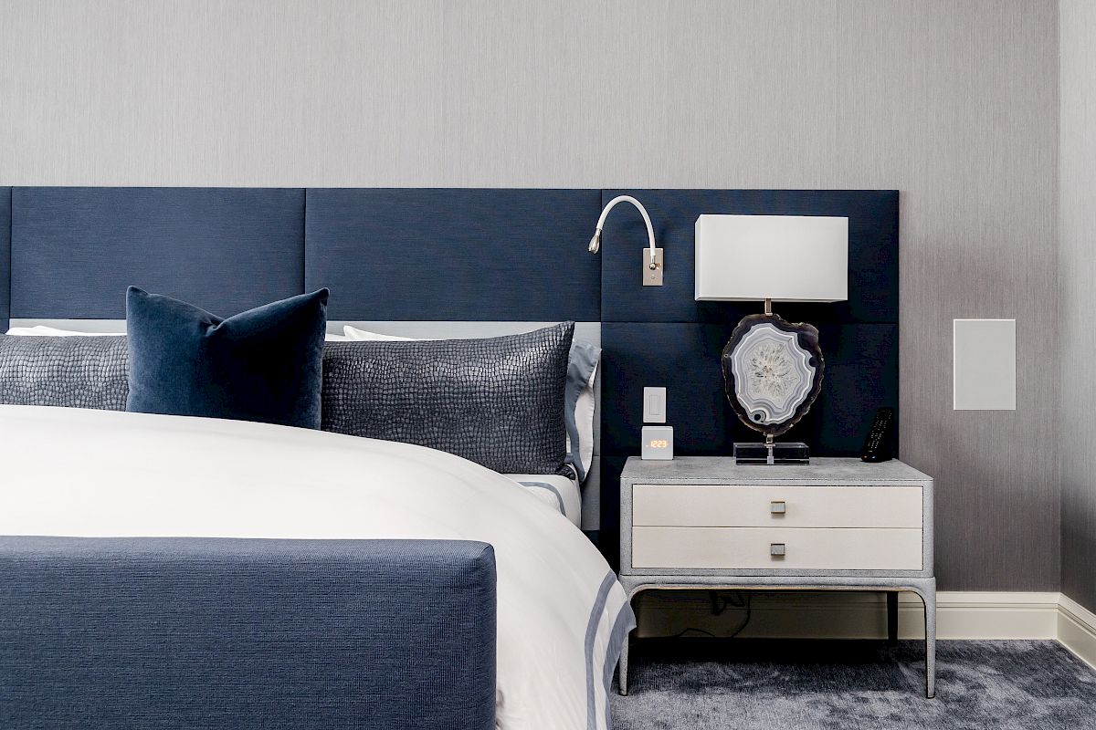 A modern bedroom with a navy blue headboard, white bedding, and a nightstand featuring a lamp and decorative item on a carpeted floor.