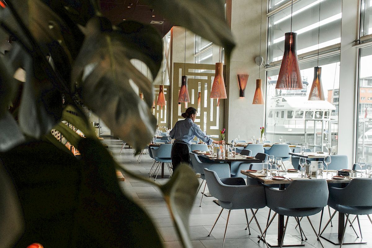 A person sets tables in a modern restaurant with stylish lighting, leafy plants, and large windows overlooking the outside.