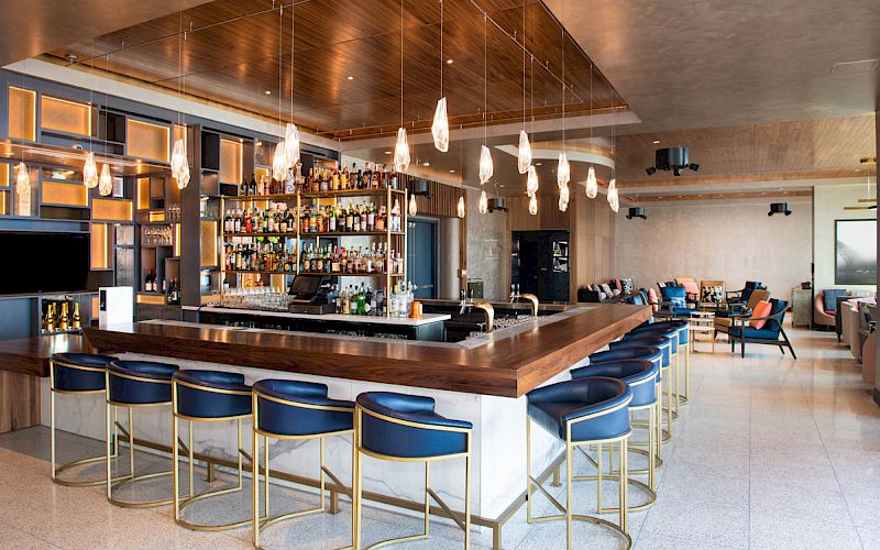 A modern bar with a central counter, blue bar stools, pendant lighting, and shelves stocked with bottles, in a chic lounge area.
