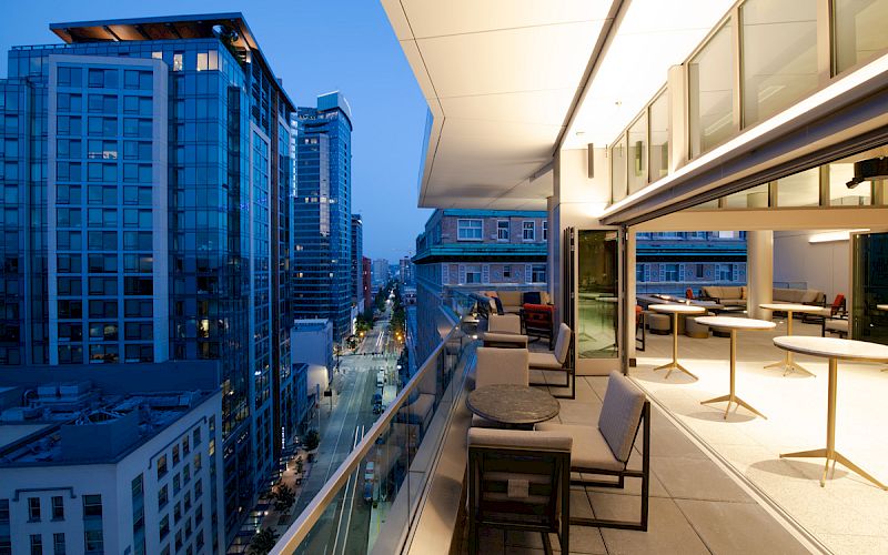 A modern cityscape at dusk with tall buildings and a sleek rooftop patio featuring tables and chairs, providing a stylish urban view.