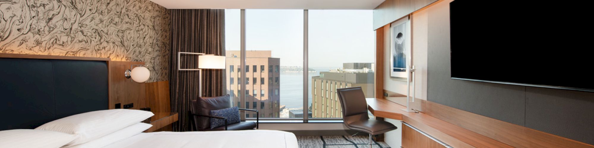 A modern hotel room with a large bed, desk, chair, and widescreen TV facing a window with a city view across several buildings.