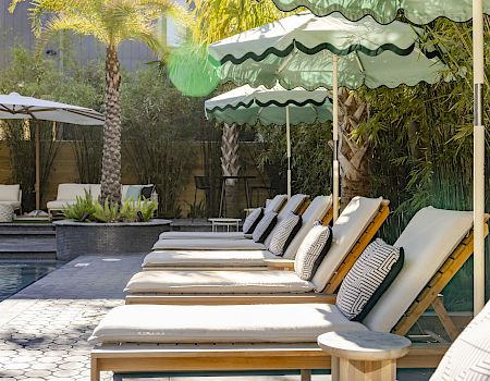 Poolside patio with white lounge beds under green striped umbrellas, pillows, and a small pool fountain area nearby.
