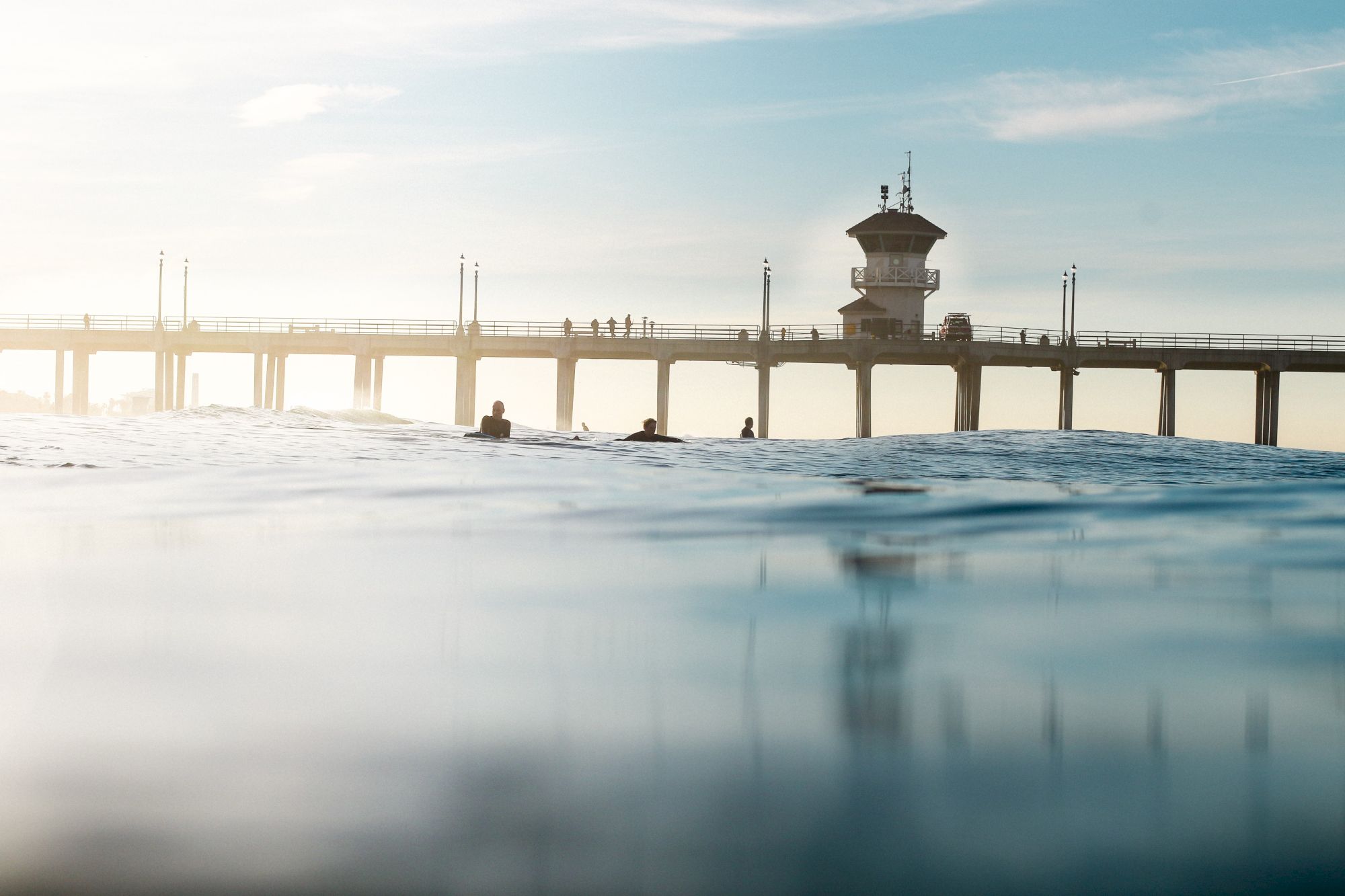 An ocean view featuring a pier with a few surfers in the water and a cloudy sky in the background.